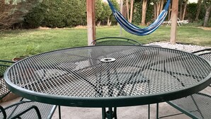 A round metal table with a mesh top, surrounded by metal chairs, set outdoors with a hammock and trees in the background.