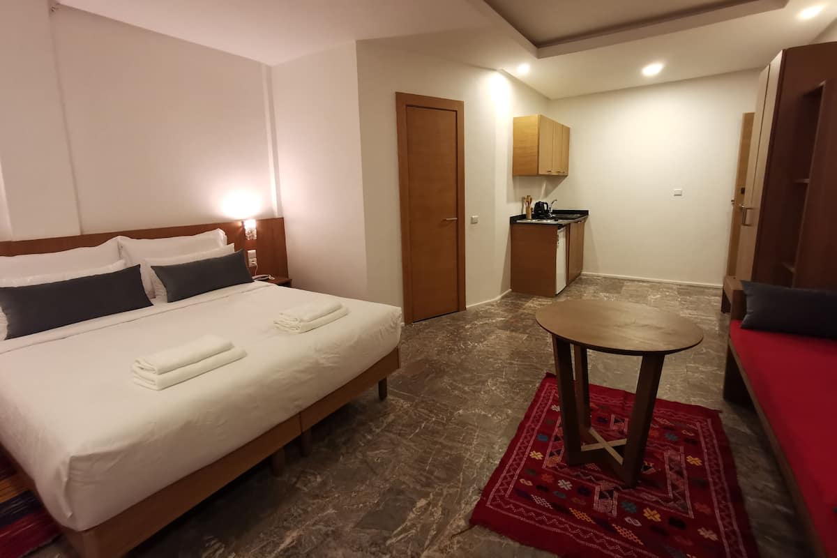 A hotel room with a bed, a small table, a red sofa, a kitchenette, and a door.