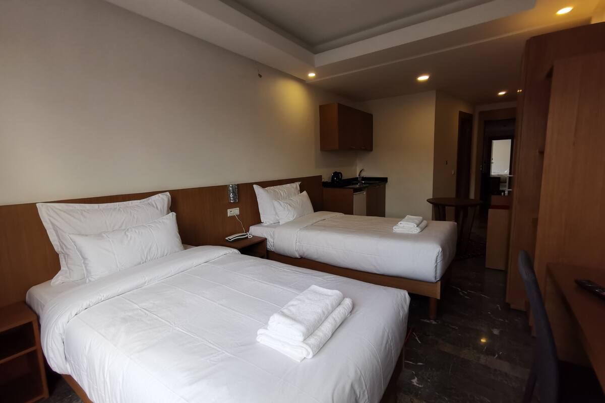 A hotel room with two beds, a small kitchenette, and a bathroom visible in the background.