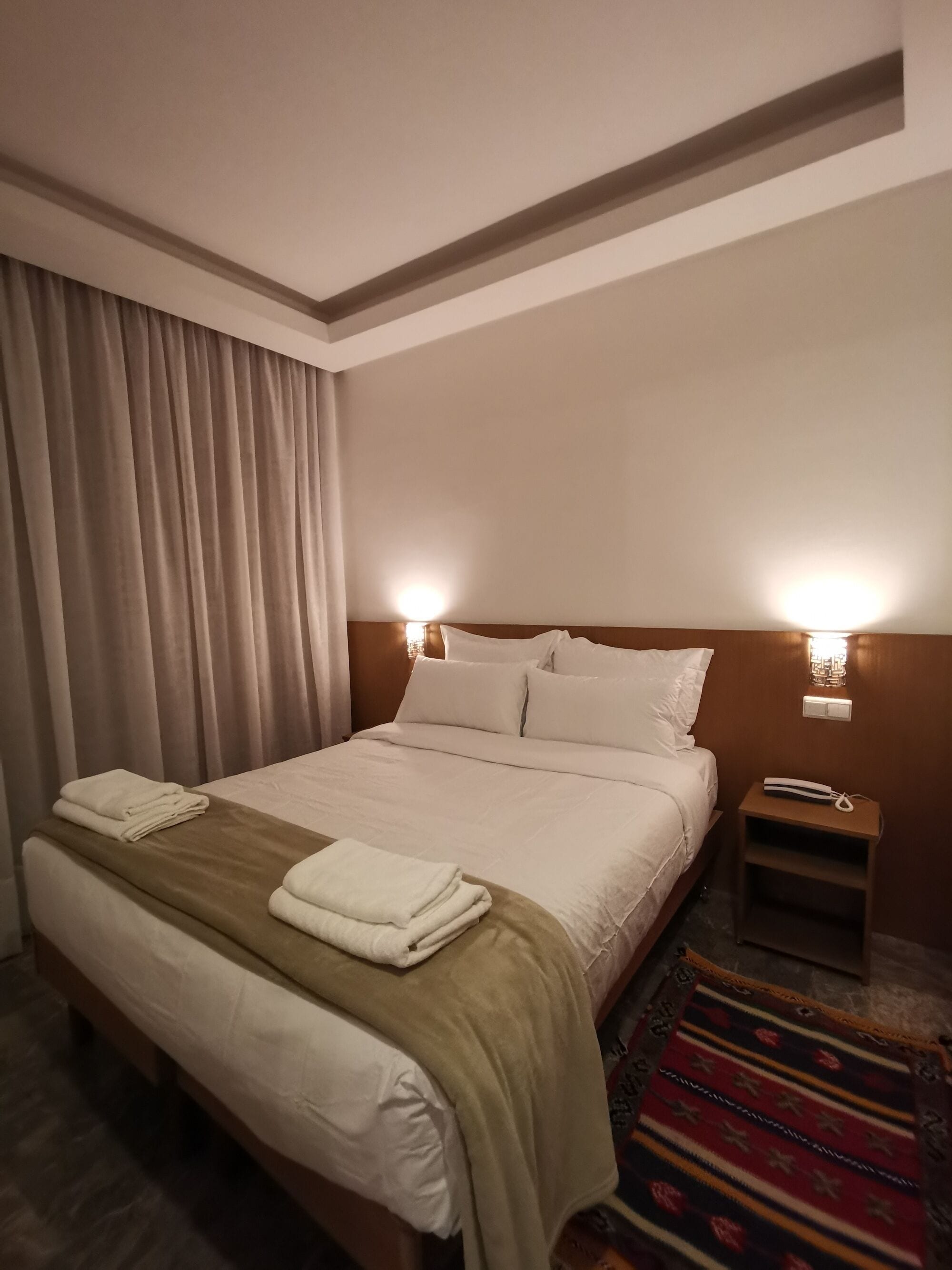 Et hotelværelse med en stor seng, to sengeborde, et natbord, en lampe og en tæppe med mønster.