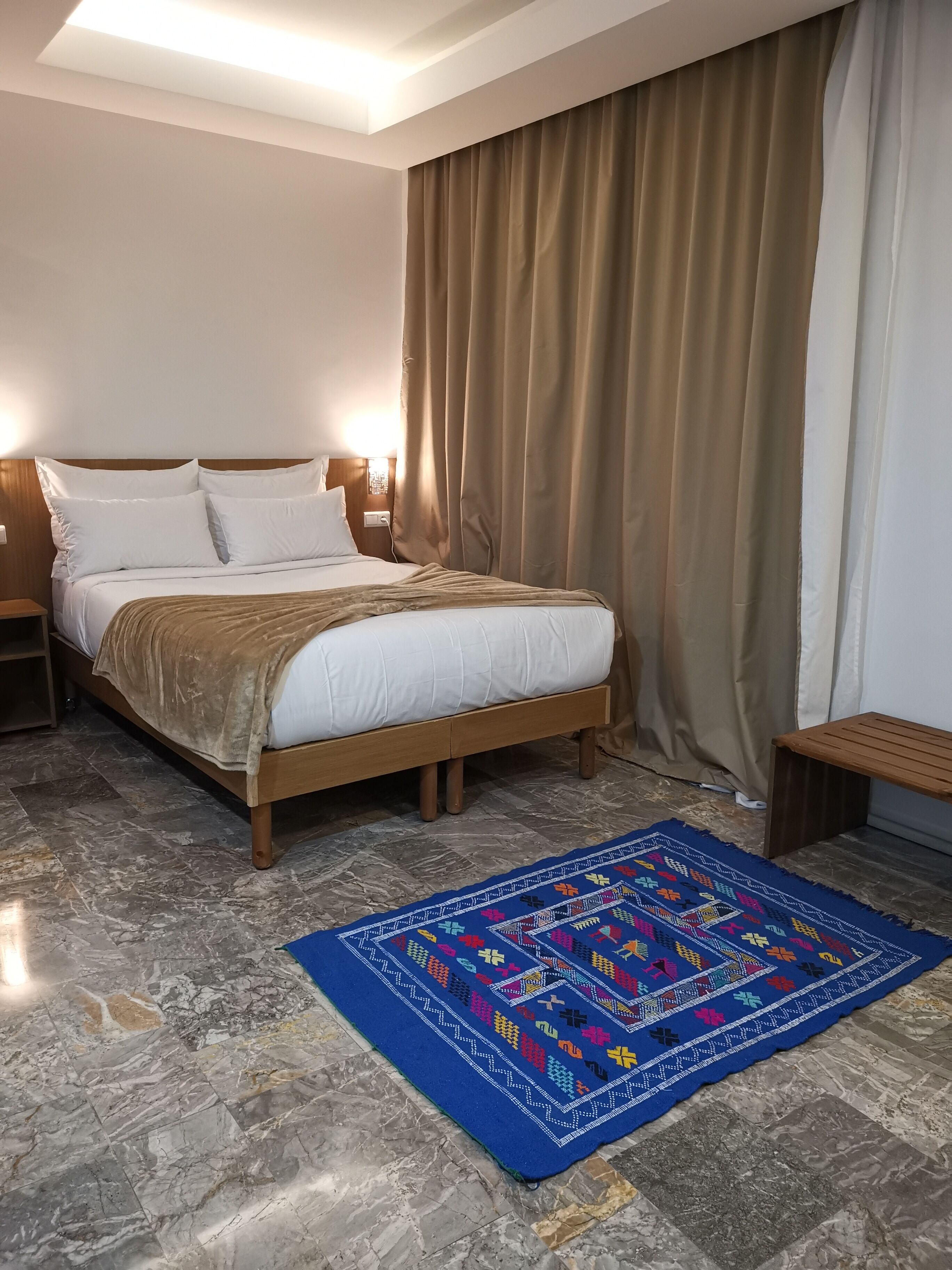 Een hotelkamer met een bed, een houten hoofdbord, beige gordijnen, een vloerkleed met een blauw patroon en een marmeren vloer.
