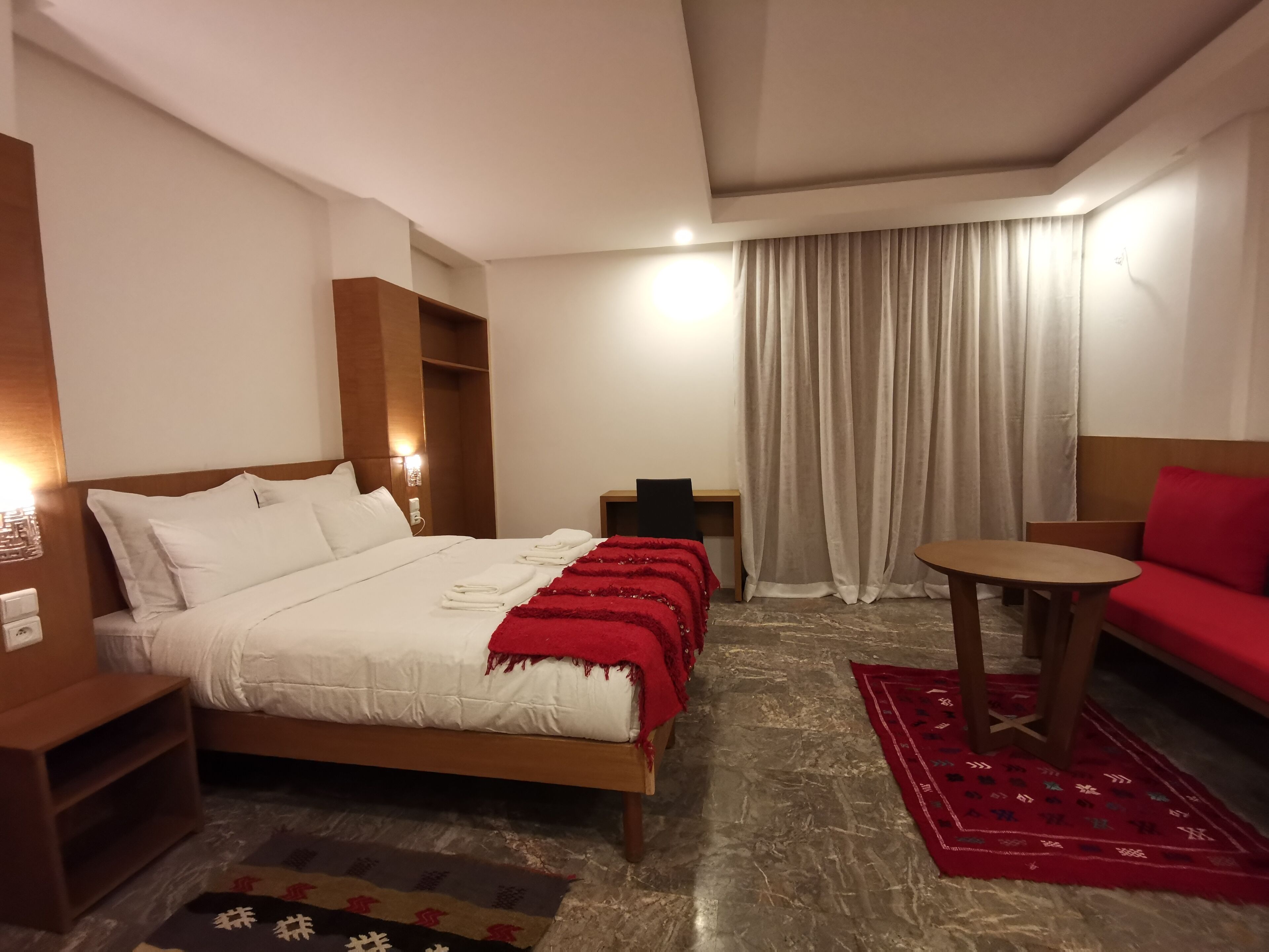 A hotel room with a bed, a small table, a red sofa, and a window with curtains.