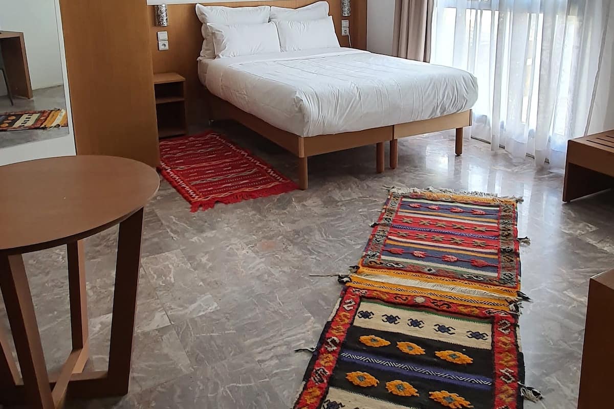 A hotel room with a bed, a small table, a red rug, and a patterned rug on the floor.