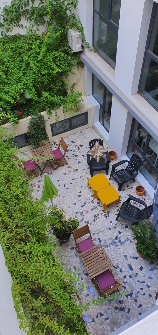 Une terrasse extérieure dotée d’un sol en mosaïque, entourée de verdure et aménagée avec des chaises en bois et en plastique, une table jaune et un petit banc en bois.