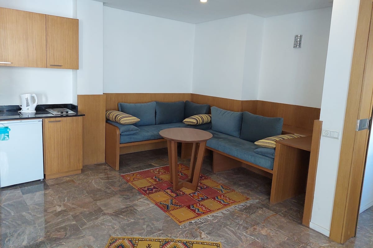 A compact living space with a kitchenette, a blue sofa with striped cushions, a small round table, and a patterned rug.
