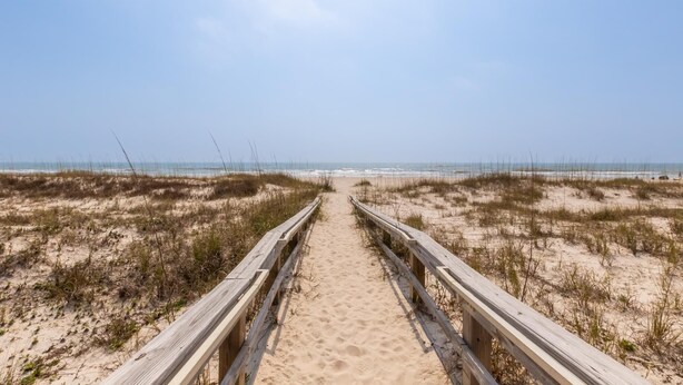 Une promenade en bois qui mène à une plage bordée de dunes et dotée d’une végétation clairsemée.