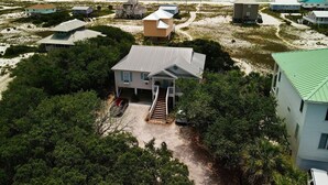 Une propriété située sur la côte, comprenant plusieurs bâtiments, dont une maison principale dotée d’un porche et d’un escalier, le tout entouré d’une végétation luxuriante et avec une plage de sable en arrière-plan.