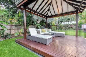 A covered outdoor seating area with a wooden deck, a white cushioned sofa, and a glass coffee table.