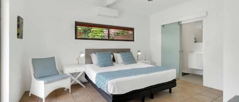 Un dormitorio con una cama, mesitas de noche, una silla, un ventilador de techo y una ventana con vistas a la vegetación.