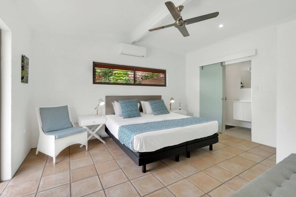 A bedroom with a bed, bedside tables, a chair, a ceiling fan, and a window with a view of greenery.