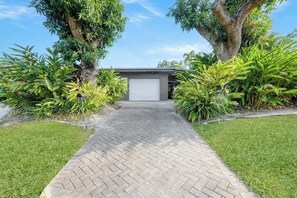 A paved driveway leading to a garage with a closed garage door, surrounded by lush greenery and a clear sky.