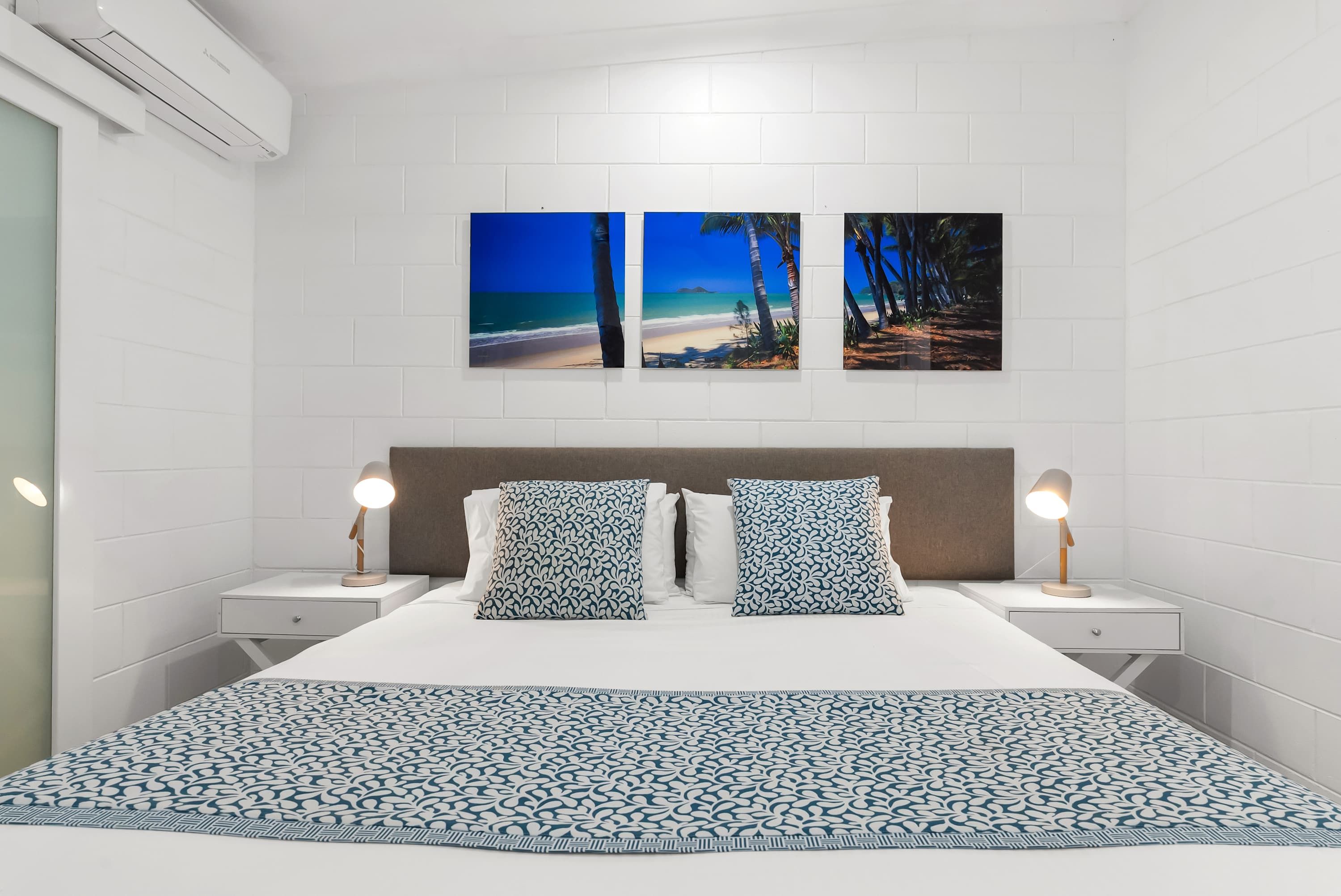 Une chambre d’hôtel comprenant un lit, deux tables de chevet, un climatiseur fixé au mur et trois tableaux encadrés au-dessus du lit.