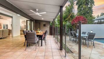 An outdoor patio with a dining table and chairs, a ceiling fan, and a view of a swimming pool and trees.