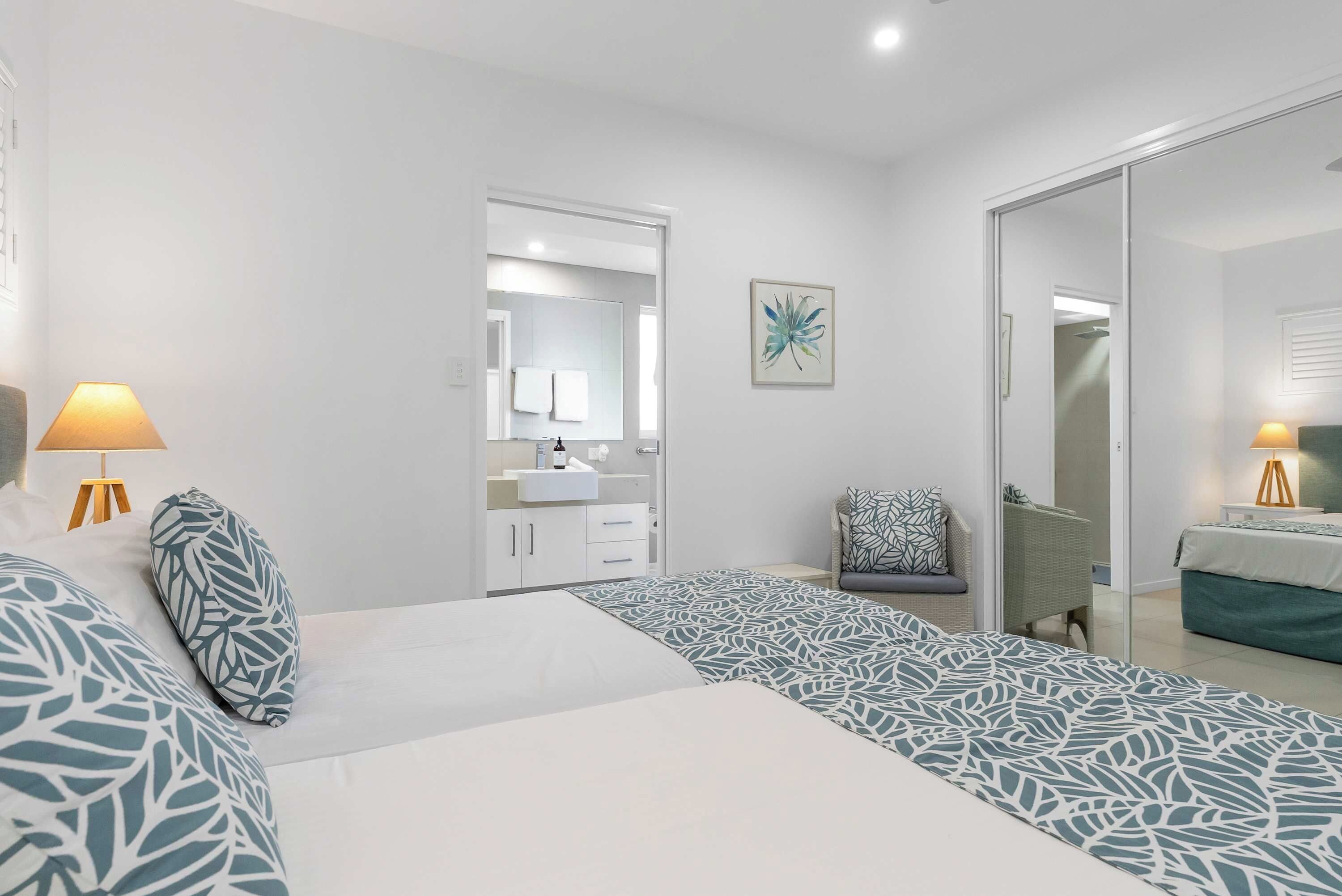 Une chambre à coucher comprenant un lit, des lampes de chevet, une chaise, un miroir de salle de bain et une œuvre d’art accrochée au mur.