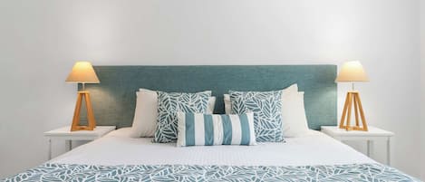 A bed with a teal headboard, white bedding, and patterned pillows. There are two bedside tables with lamps.