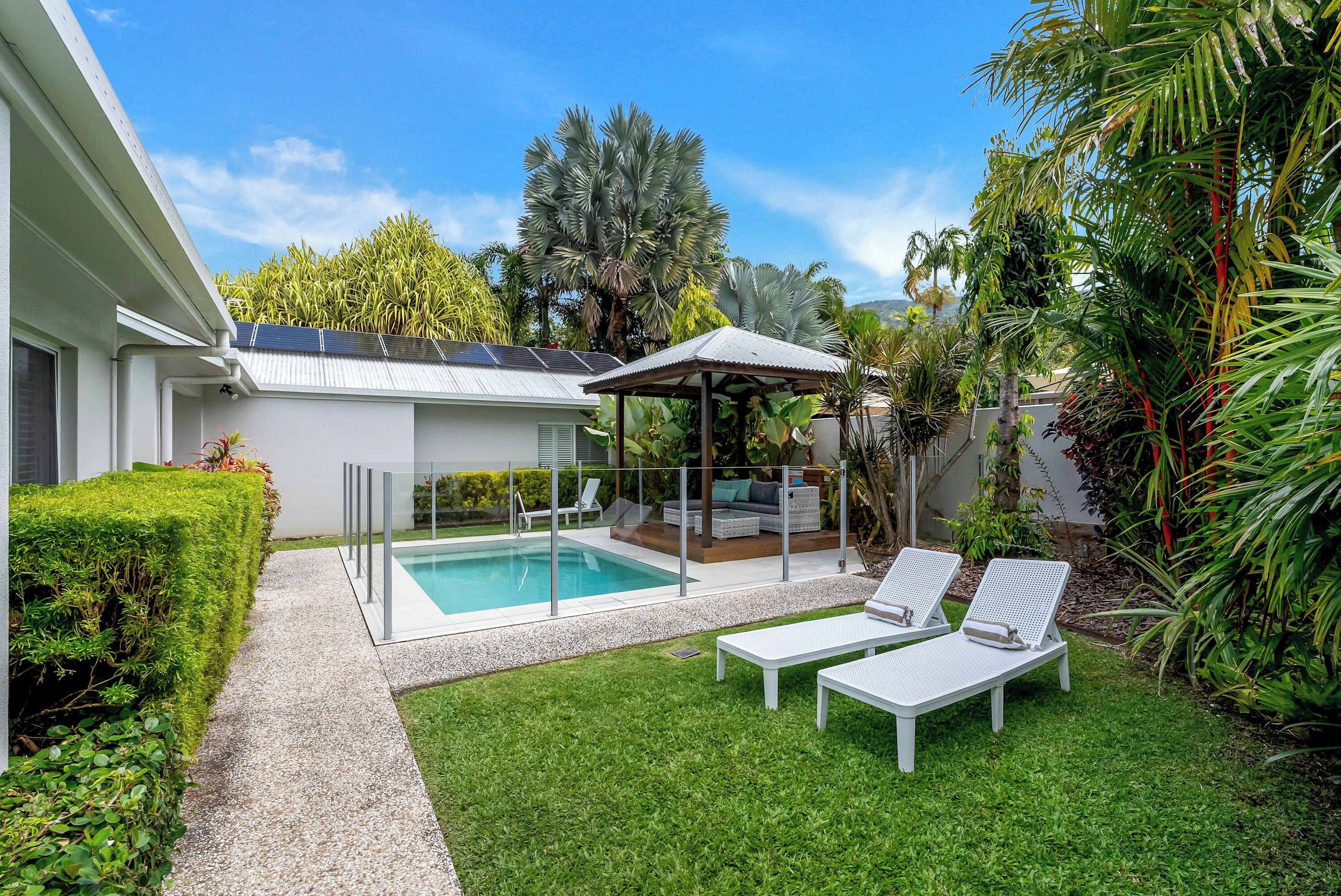 A modern outdoor pool area with a glass enclosure, two lounge chairs, and a covered seating area.