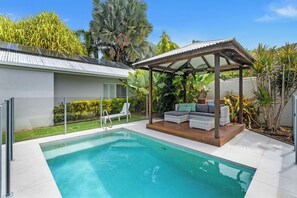 A swimming pool with a covered seating area, surrounded by lush greenery and a modern house. - The Last Resort - Villa 1 (Palm Cove)