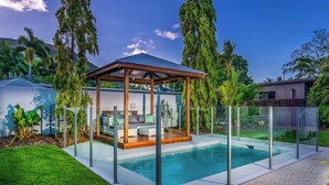 Un espace piscine extérieur moderne, doté d’une structure en verre, d’un gazebo en bois et d’une végétation luxuriante.