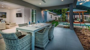 Un espace extérieur aménagé pour les repas, avec des chaises et une table en osier, un ventilateur de plafond et une vue sur une piscine et des montagnes.