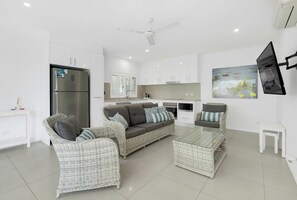 A modern living room with a sofa, coffee table, and chairs, a kitchen with white cabinets, and a ceiling fan.