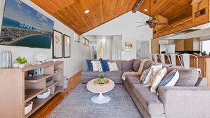 A living room with a wooden ceiling, a large TV displaying 'SAN DIEGO,' a sofa, a coffee table, and a bookshelf.