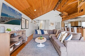 Una sala de estar con techo de madera, un televisor grande mostrando 'SAN DIEGO', un sofá, una mesita de centro y una estantería.