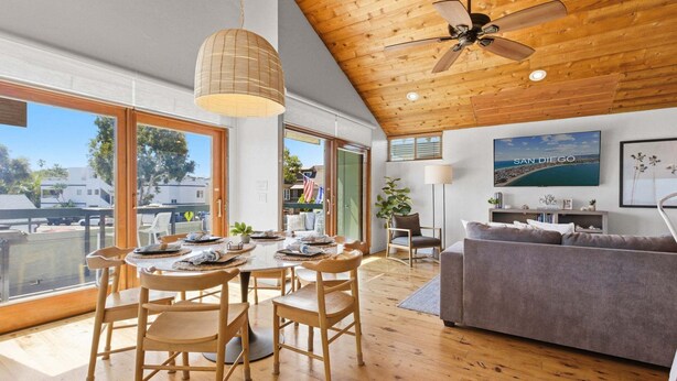A spacious living area with a dining table, wooden flooring, a ceiling fan, and a large window offering a view of the outdoors.