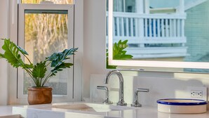 Un baño con lavamanos blanco, una planta en maceta y una ventana que da a un porche blanco.