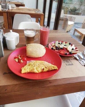 Une table sur laquelle se trouve une assiette rouge contenant une part de pizza, un petit pain rond et une salade composée de tomates, de concombres et d’olives. On aperçoit également un verre de bière et un verre de smoothie rose sur la table.