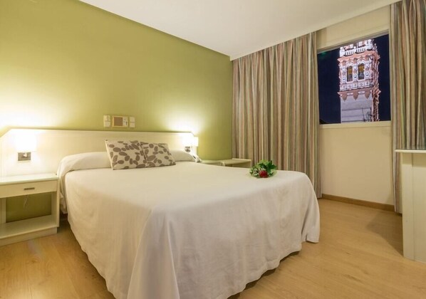 Habitación de hotel con una cama grande, una mesita de noche, una lámpara de noche, una ventana con cortinas y un adorno de pared.