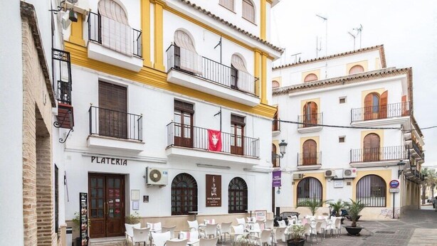 Un patio con mesas al aire libre, un edificio identificado como "PLATERIA" y una bandera roja en el balcón.