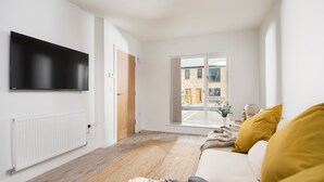 Ein modernes Wohnzimmer mit Flachbildfernseher, einer gelb gepolsterten Couch, einer Holztür und Blick auf ein Backsteingebäude durch ein großes Fenster.
