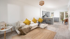 Ein modernes Wohnzimmer mit einem weißen Sofa, gelben Kissen, einem Holztisch und einer Küche im Hintergrund.