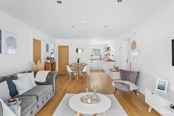 A modern living room with a grey sofa, a dining table, and a kitchen in the background. - Plover 1, The Cove - On site pool - Wi-Fi (Brixham)