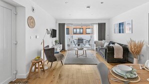 A modern living room with a sofa, a dining table, and a kitchen area visible through an open door. - Plover 1, The Cove - On site pool - Wi-Fi (Brixham)