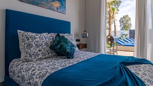 Een slaapkamer met een blauw hoofdeinde, beddengoed met een patroon, een schilderij aan de muur en uitzicht op een zwembadgedeelte met ligbedden.