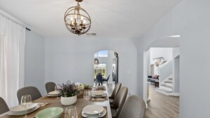 Un espace repas avec une longue table dressée pour six personnes, un lustre et une vue sur une autre pièce.