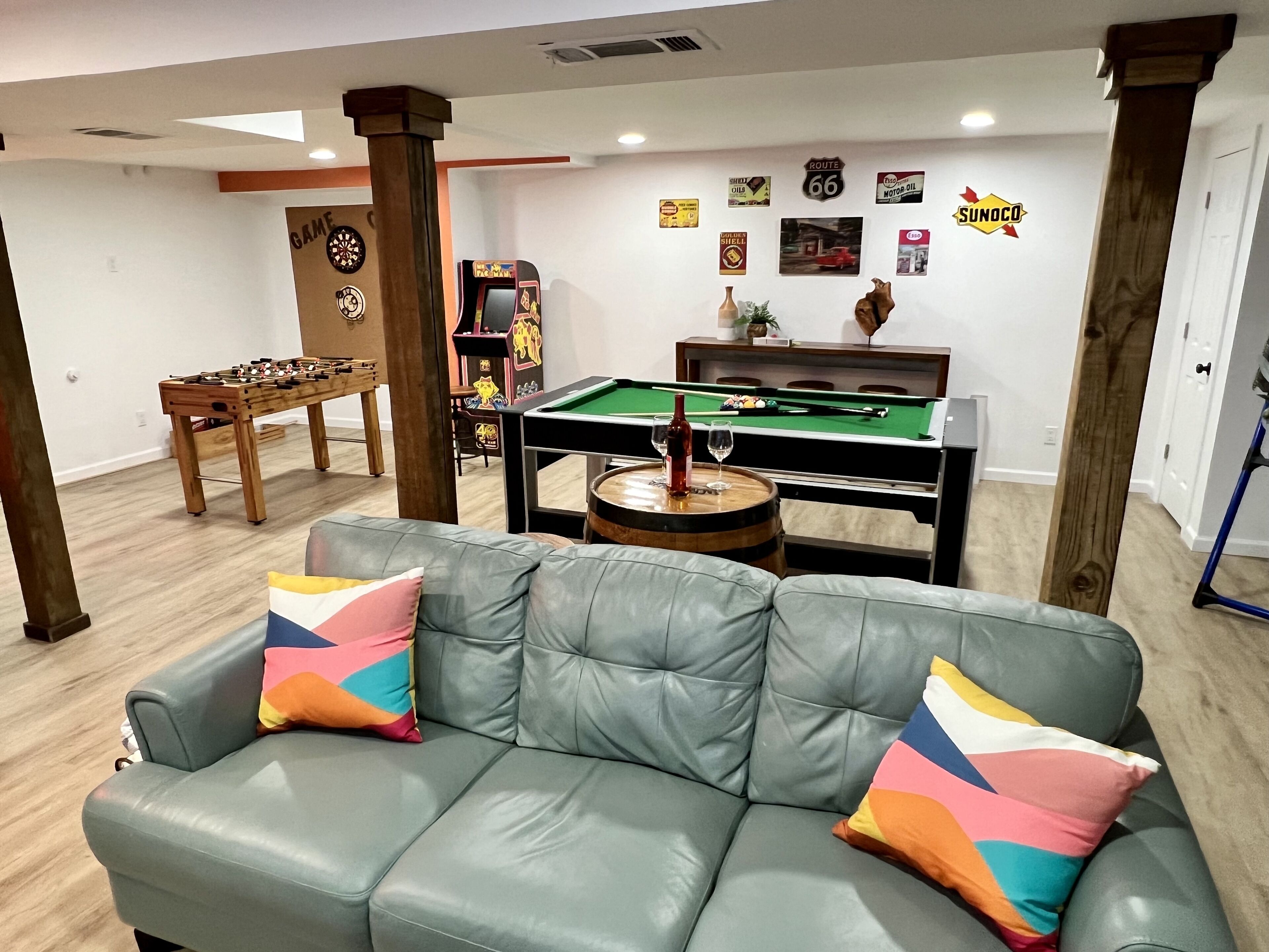 A room with a green leather sofa, a foosball table, a pool table, and various wall decorations including Route 66 signs and a Sunoco sign.
