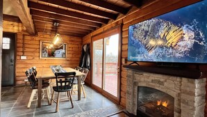 Interior acolhedor de cabana com mesa de jantar de madeira, cadeiras, lareira e uma TV grande na parede.