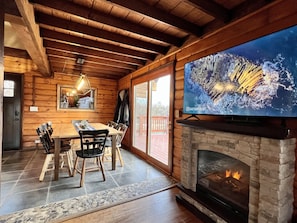 Interior acogedor de una cabaña con una mesa de madera, sillas, chimenea y un televisor grande en la pared.
