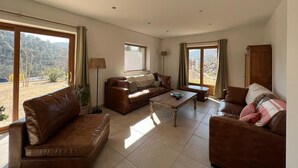 Een ruime woonkamer met een bruine leren bank, een houten salontafel en uitzicht op het landschap door grote ramen.