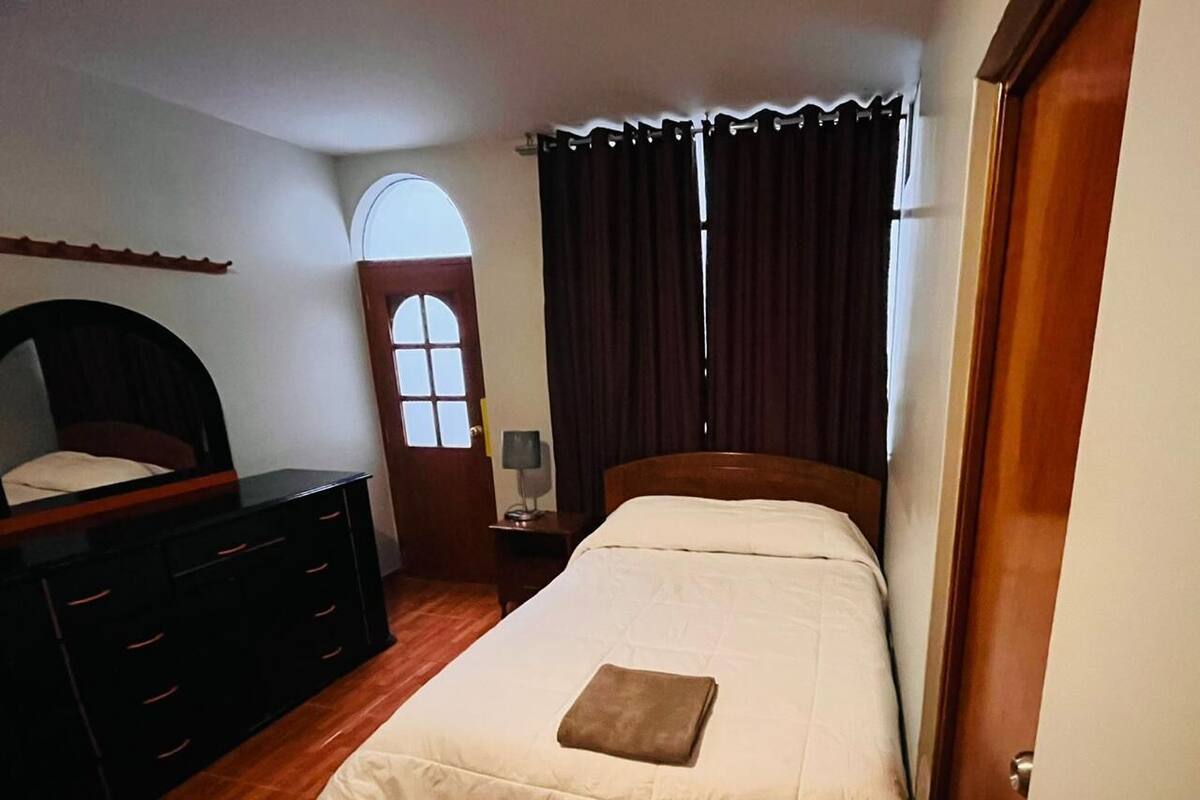 A bedroom with a bed, a dresser, a nightstand, a lamp, and a window with curtains.