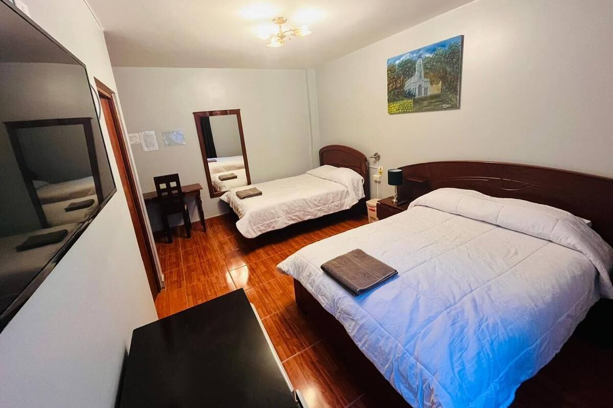 A hotel room with two single beds, a wooden floor, a desk with a chair, a mirror, and a painting on the wall.