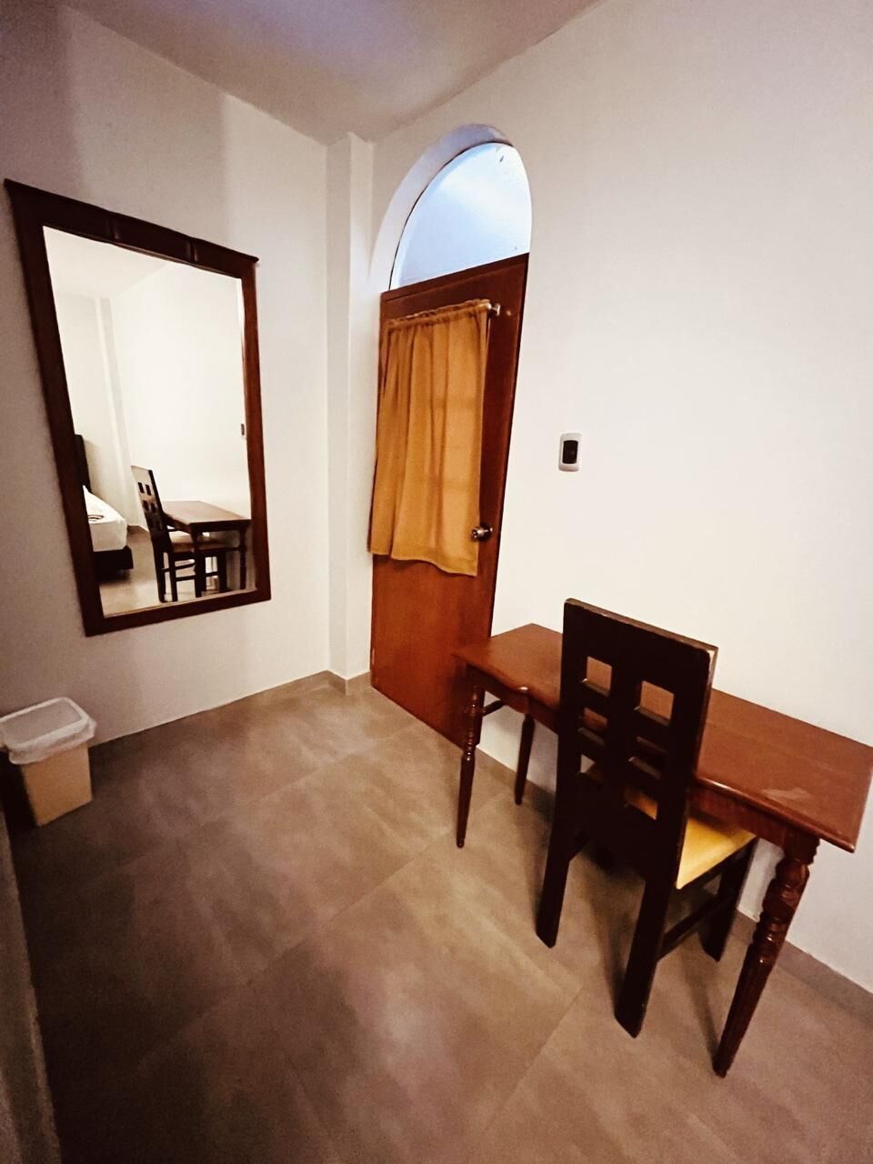 Una habitación con un escritorio de madera, una silla, un espejo, una puerta con cortina y un pequeño cubo de basura.