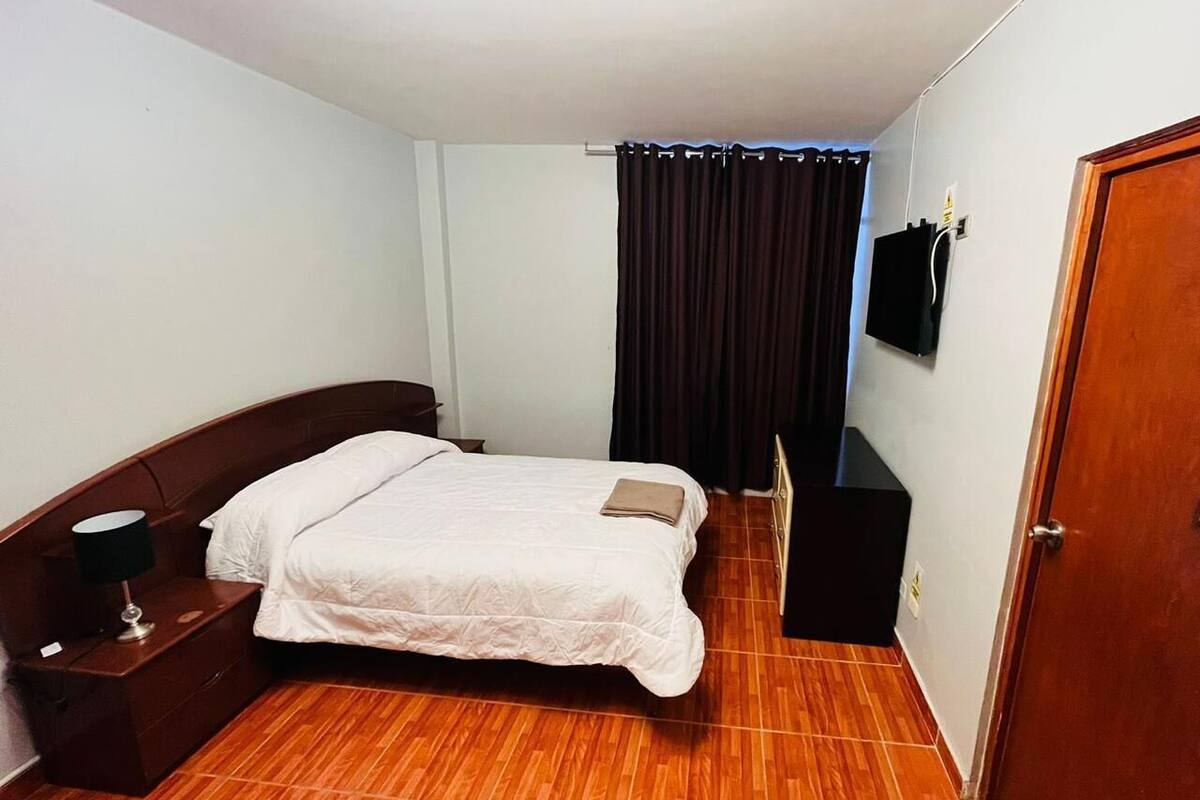 A bedroom with a wooden floor, a bed with white bedding, a nightstand with a lamp, a TV on the wall, and a door.