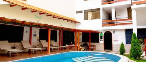 A swimming pool with a tiled edge, a covered seating area with chairs, and a multi-story building with balconies.