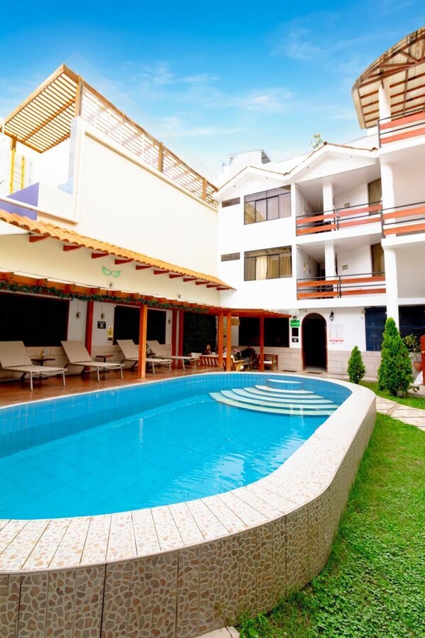 A swimming pool with a tiled edge, a covered seating area with chairs, and a multi-story building with balconies.