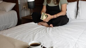Une chambre d’hôtel avec un lit, une table de chevet avec une lampe, un plateau avec des pâtisseries et du café, et un tableau au mur.