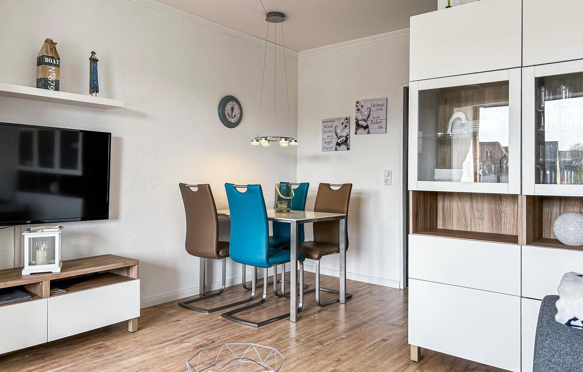 Una sala de estar moderna con una televisión de pantalla plana, una mesa de comedor con cuatro sillas, una estantería y un reloj de pared decorativo.