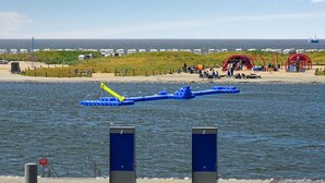 Een kustgebied met een pier die in het water uitloopt, een zandstrand met mensen en een uitzicht op zee in de verte.